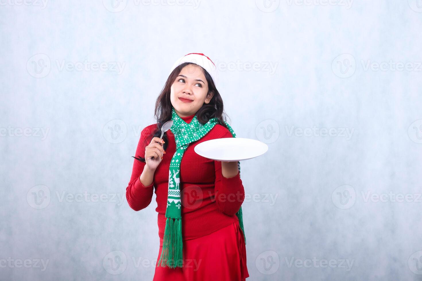 expression of young girl wearing red sweater red sweater with santa hat and scarf celebrating merry christmas and new year, smile get idea carry white plate and hold spoon, isolated white background photo