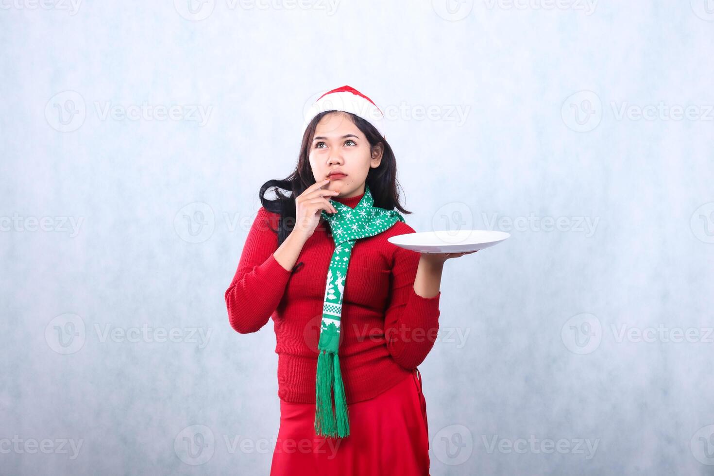 gesture of young Asian woman wearing red sweater with Santa hat and scarf, thinking hands raise white plate and hold chin, celebrating merry christmas and new year isolated on white background photo