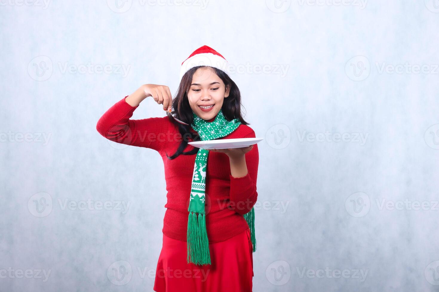 Charming young Asian woman wearing red sweater with Santa hat and scarf, smiling to food, carrying white plate and holding spoon, celebrating merry christmas and new year isolated on white background photo