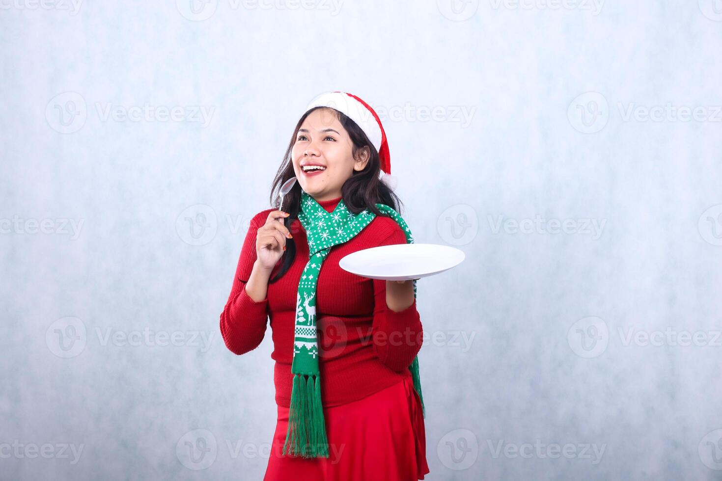 Charming young Asian woman wearing red sweater with Santa hat and scarf, cheerful while thinking carrying white plate and holding spoon, celebrating merry christmas isolated on white background photo