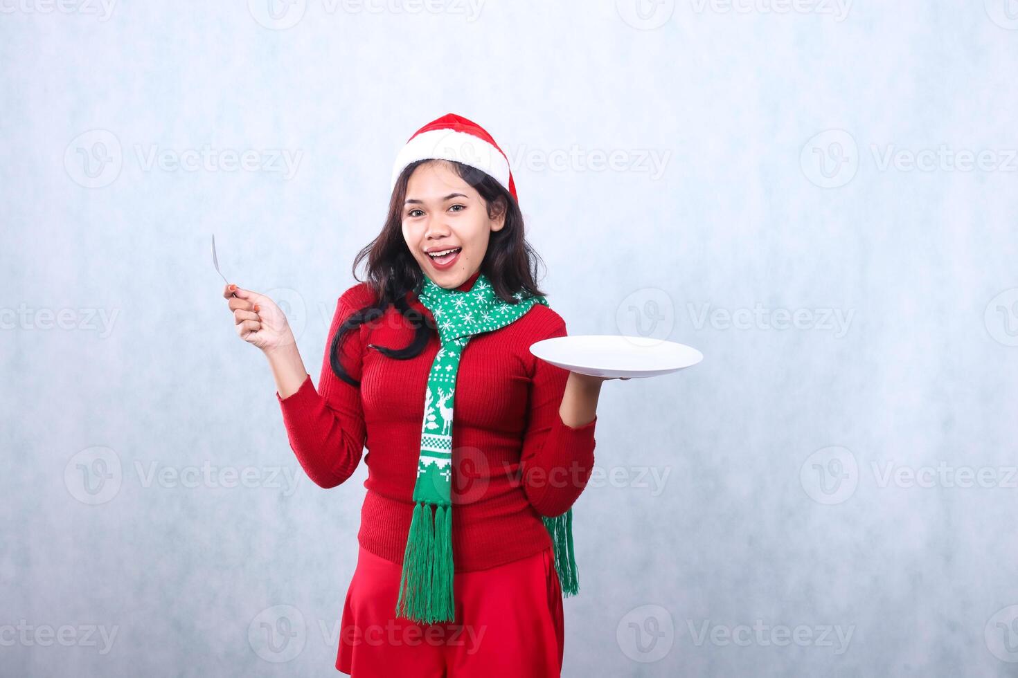 Charming Asian girl wearing red sweater with Santa hat and scarf, cheerful to food carry white plate and hold spoon, celebrating merry christmas and new year isolated on white background photo