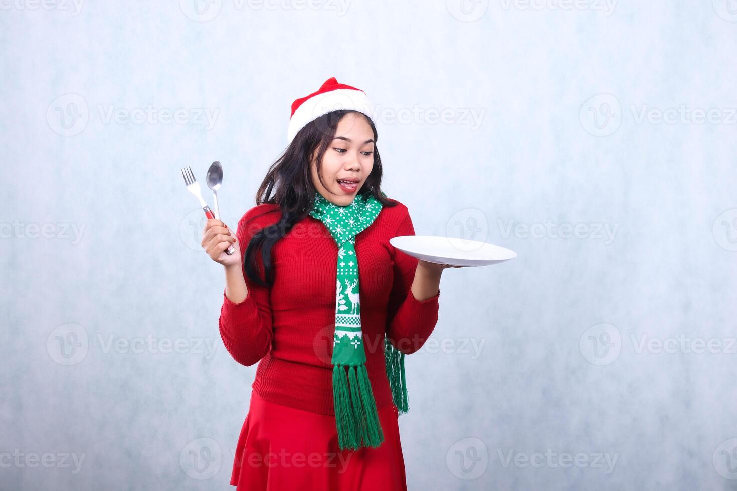 gesture young asian girl wearing red sweater with Santa hat and scarf, tempted to see food carry white plate and hold spoon fork, celebrate merry christmas and new year isolated on white background photo