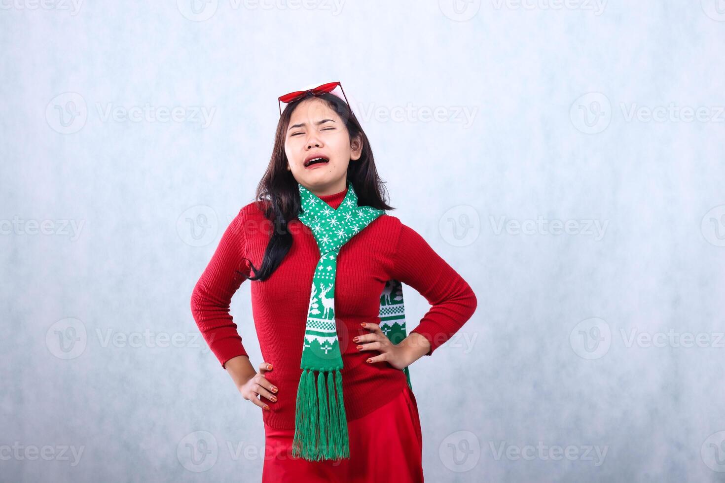 expression of young asian woman wearing red sweater with Santa hat and scarf celebrating merry christmas and new year, crying sobbing eyes closed, wearing glasses on head, isolated on white background photo