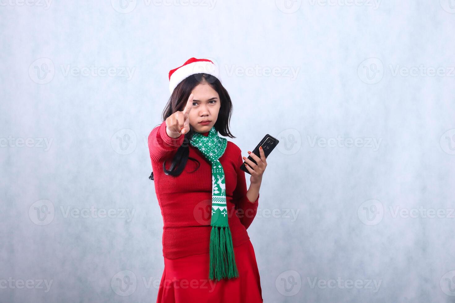 young woman serious to camera hand holding mobile phone and reject sign, wearing red sweater with Santa hat and scarf celebrating merry christmas and new year, isolated on white background photo