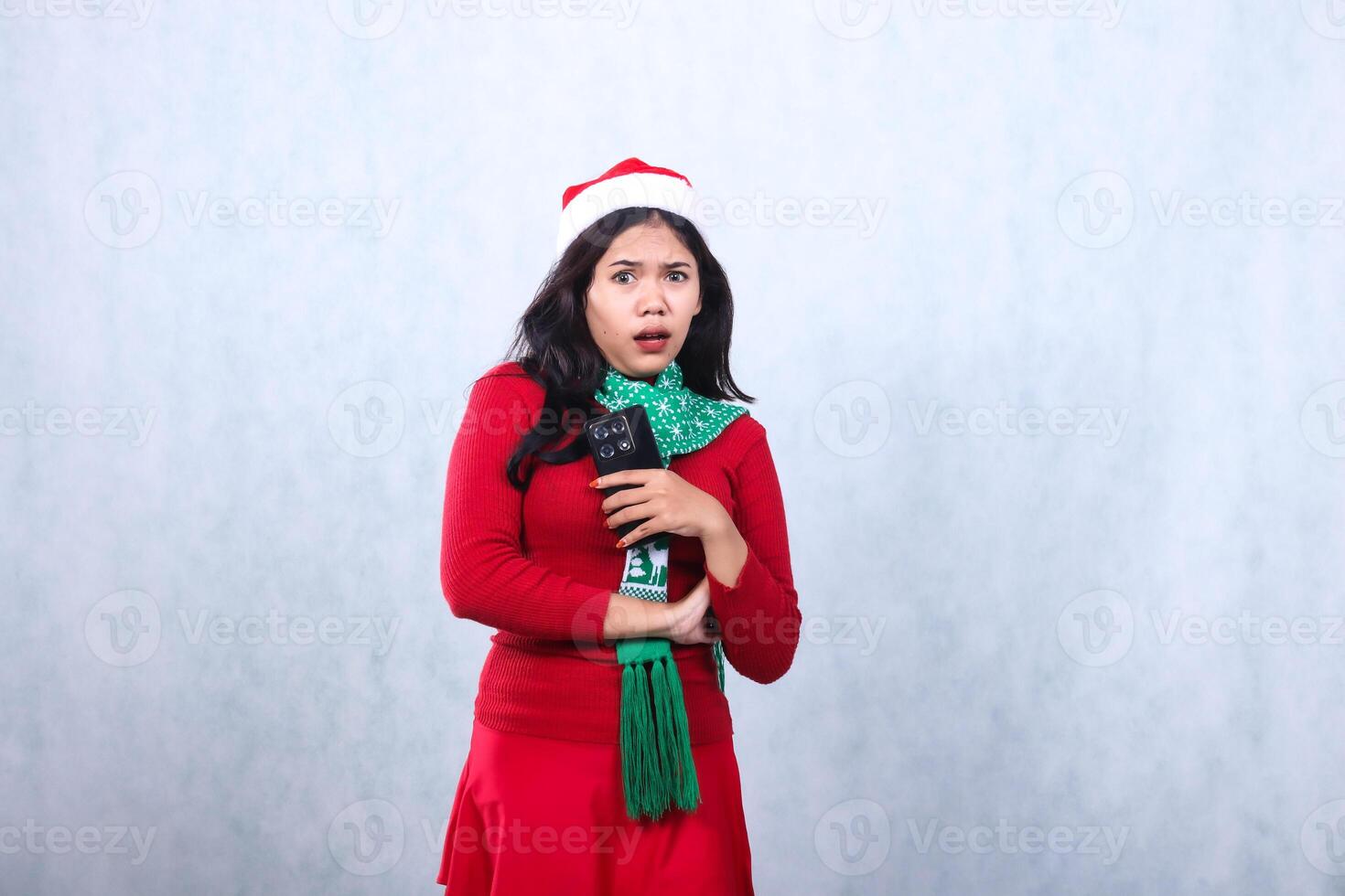 portrait of Asian girl wearing christmas sweater, Santa hat and scarf, scared shocked to camera, hands together while holding cellphone mobile phone, isolated on white background photo