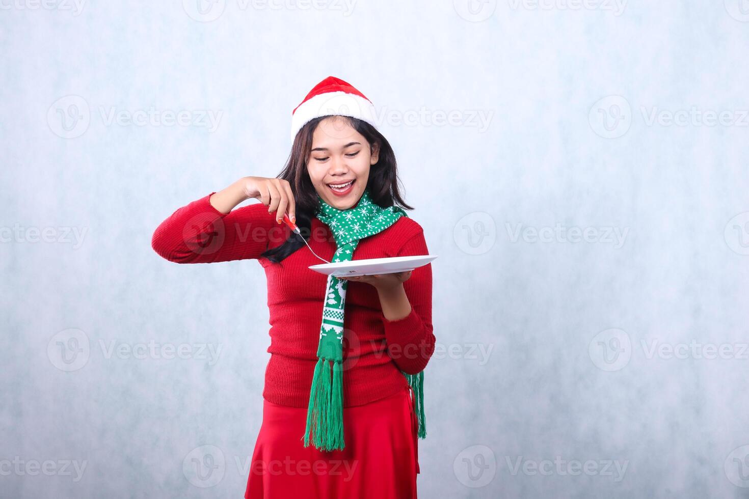 charming young woman wearing red sweater with Santa hat and scarf, cheerful to the plate carrying white plate and scooping food with fork, celebrating merry christmas isolated on white background photo
