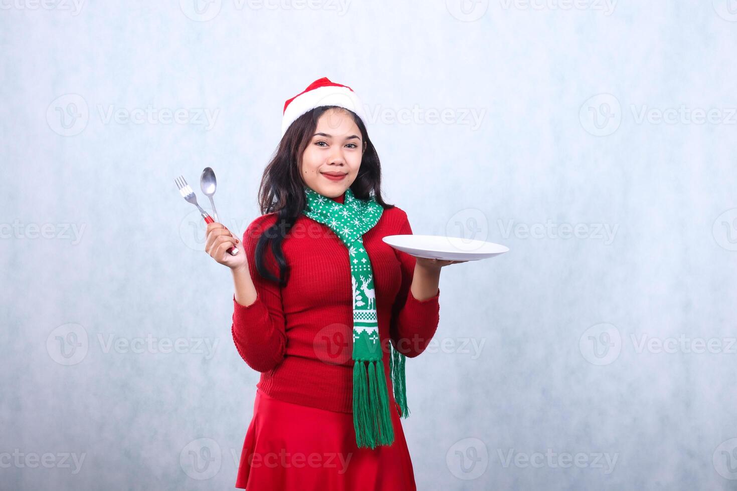 Charming adult woman wearing red sweater with Santa hat and scarf, graceful smile looking forward, carry white plate and hold spoon fork, celebrate merry christmas isolated on white background photo