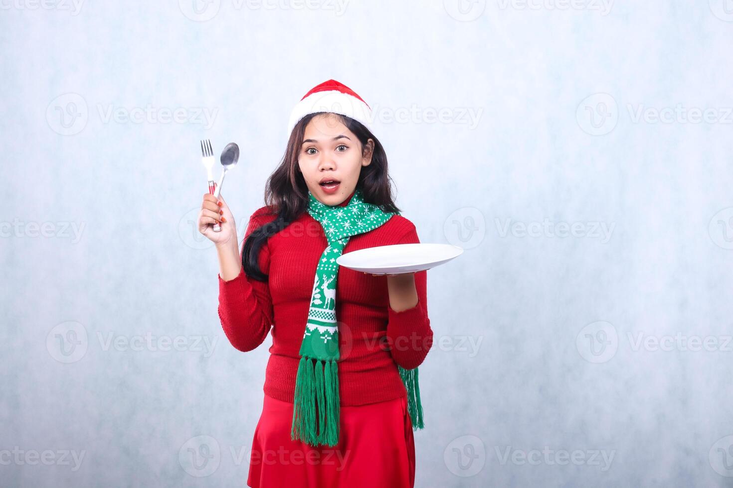 Charming young woman wearing red sweater with Santa hat and scarf, surprised to camera carrying white plate and holding spoon fork, celebrating merry christmas isolated on white background photo