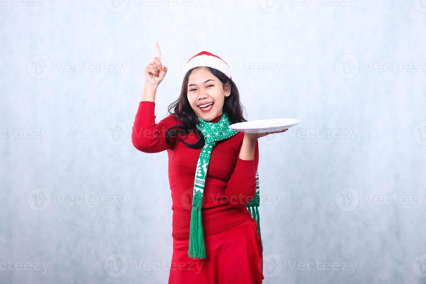 portrait of woman wearing red sweater red sweater with santa hat and scarf celebrating merry christmas,cheerful looking at camera hand lifting white plate and pointing up ,isolated on white background photo