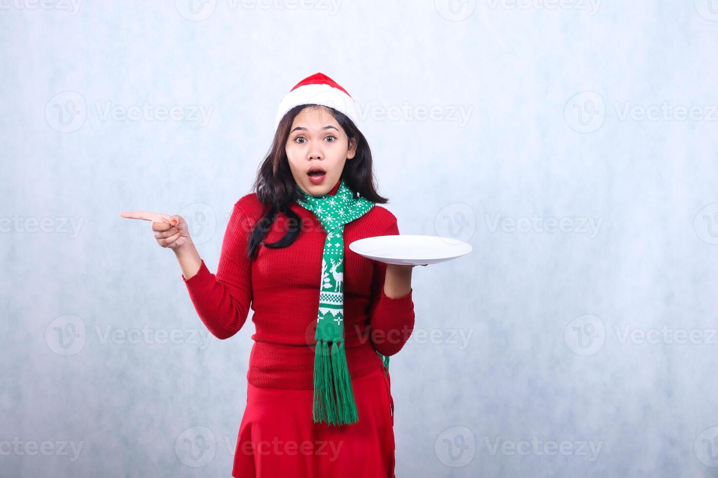 woman wearing red sweater with Santa hat and scarf, surprised to camera holding white plate while pointing right, celebrating merry christmas and happy new year isolated on white background photo