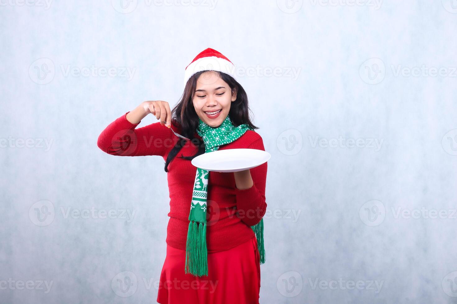 Charming indonesia woman wearing red sweater with Santa hat and scarf, cheerful to the plate carrying white plate and scooping with spoon, celebrating merry christmas isolated on white background photo