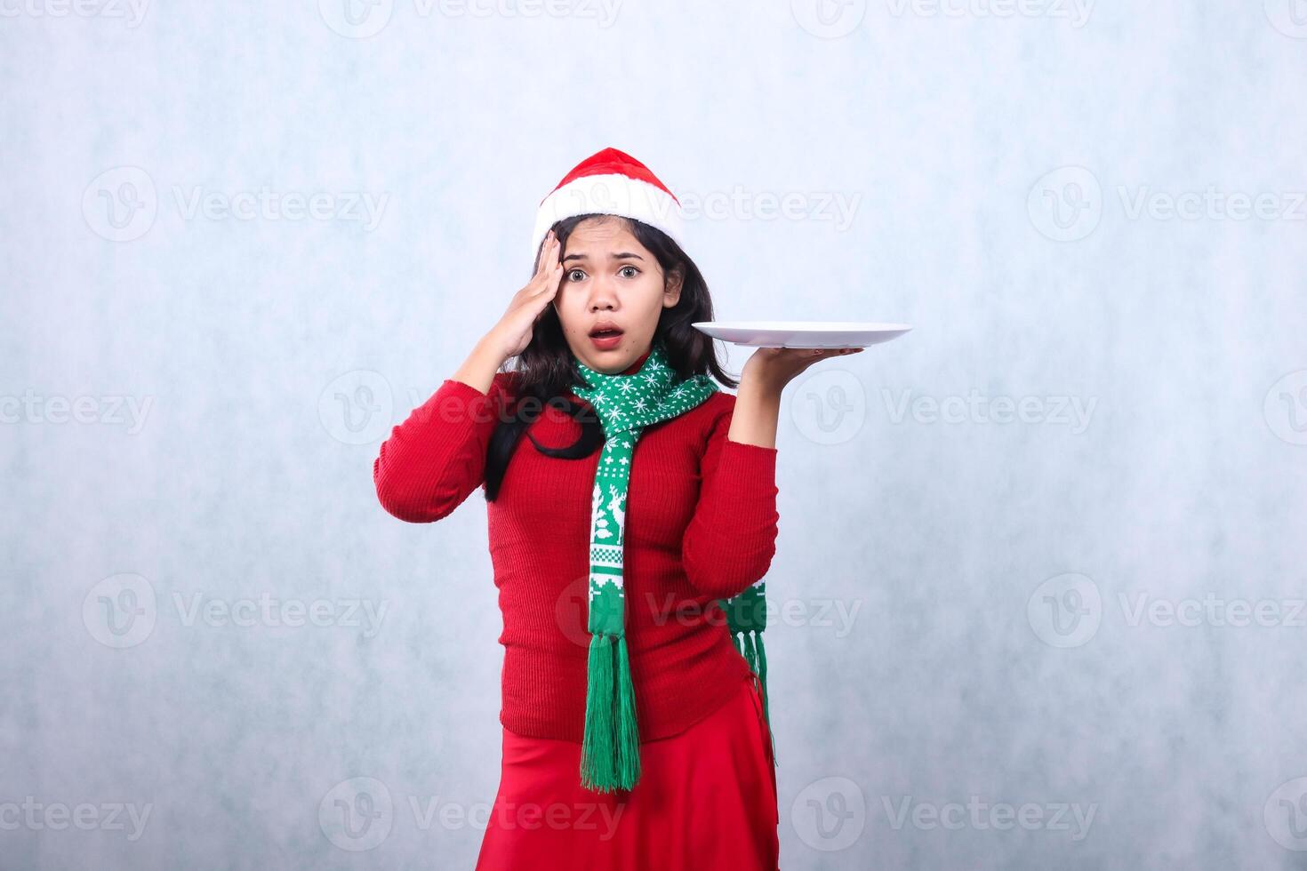 Charming woman wearing red sweater with Santa hat and scarf, shocked looking at camera, hand lifting white plate and holding forehead, celebrating merry christmas isolated on white background photo