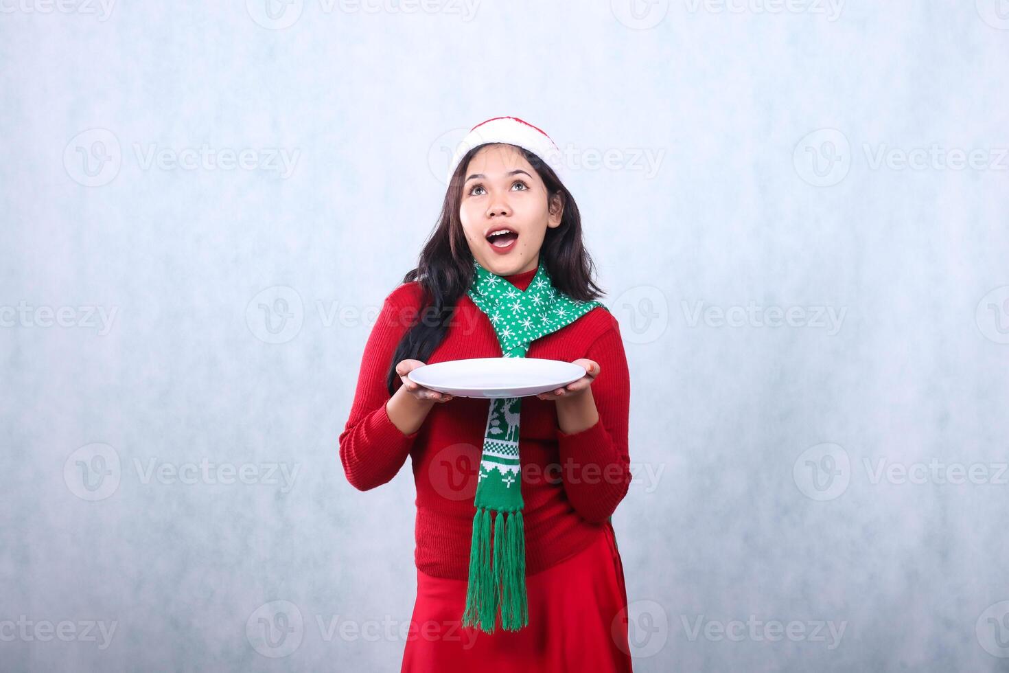 expression of young asian woman wearing red sweater red sweater with santa hat and scarf celebrating merry christmas, candid surprise on both hands carrying white plate, isolated on white background photo