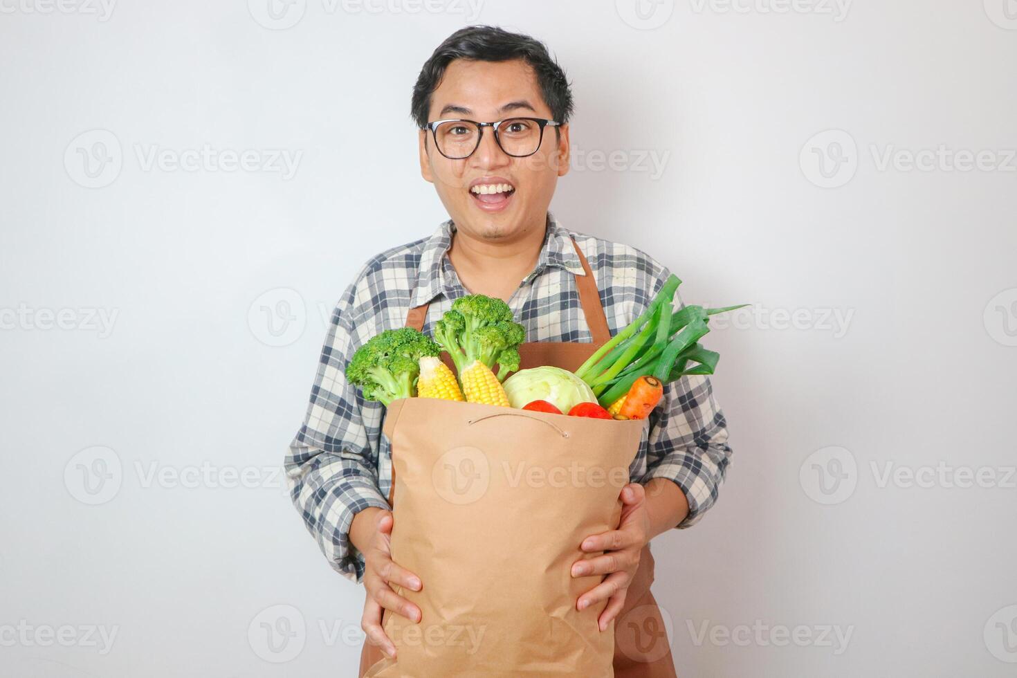 Young smiling happy asian man wear shirt and apron hold brown craft bag for takeaway mock up with food products show thumb up gesture isolated on plain blue background. photo