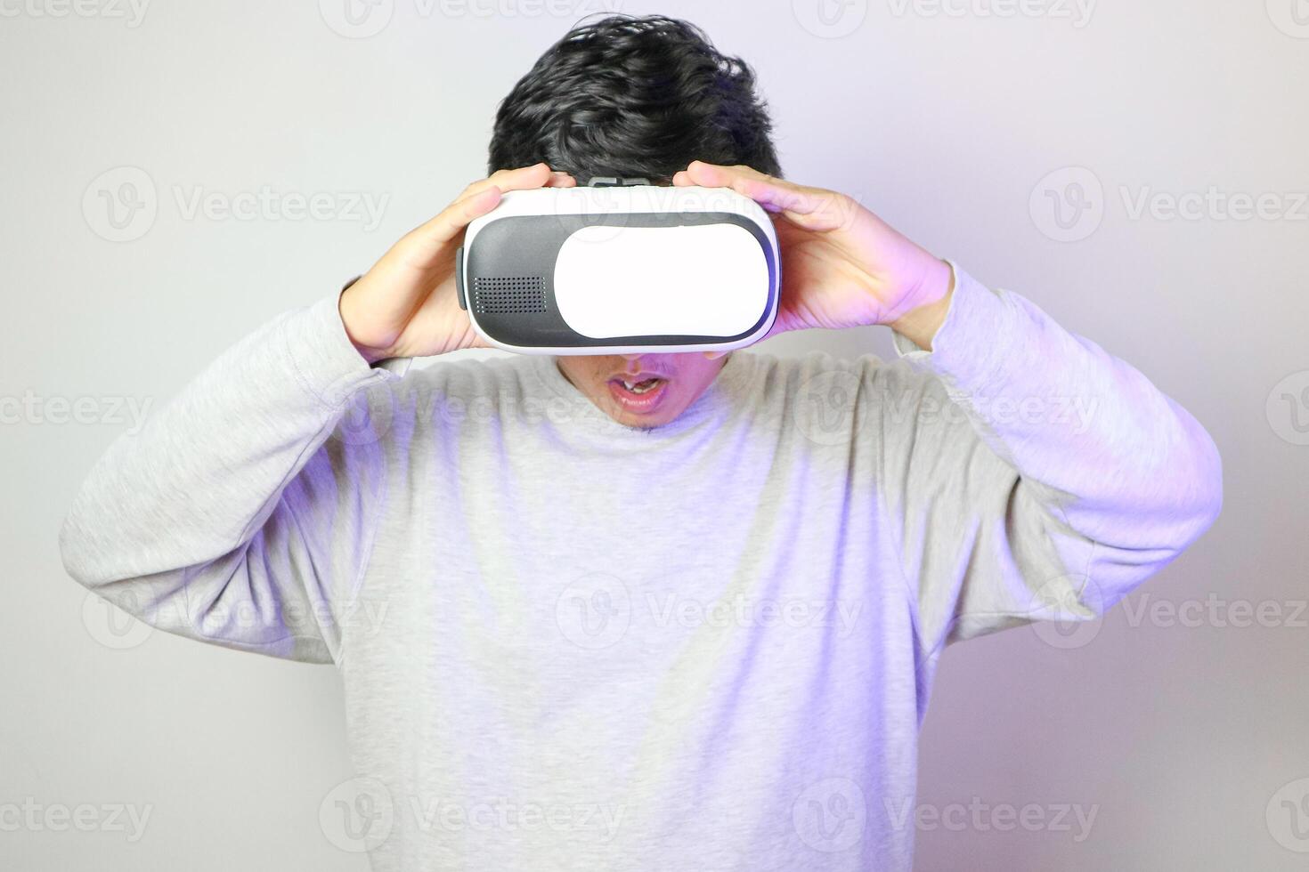 Successful Asian male virtual reality gamer taking off vr headset and looking around at studio, isolated portrait over dark blue background with double coloured lightning photo