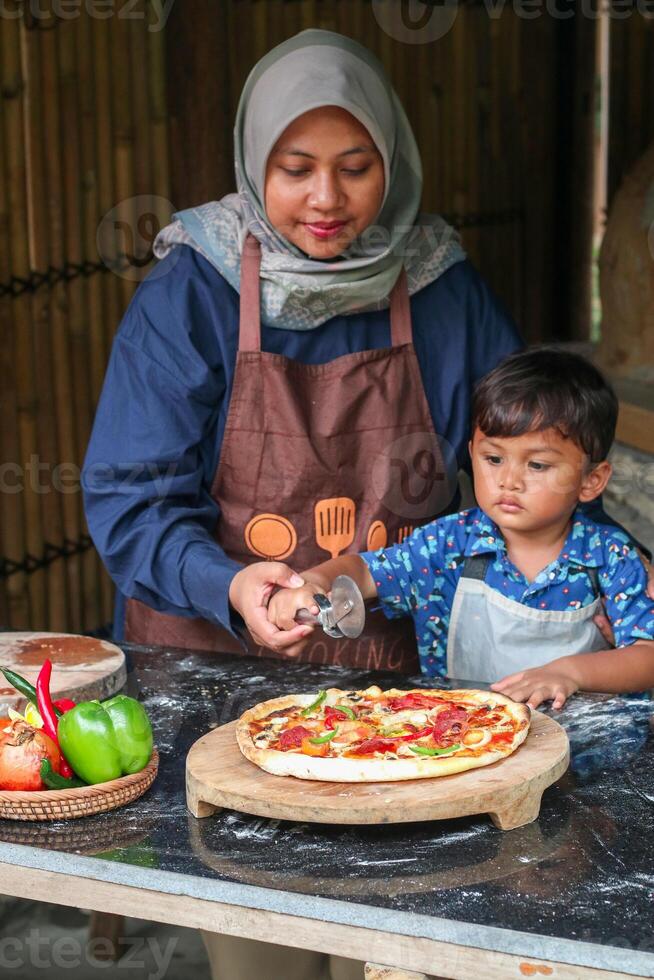 Muslim Mother And Little son Rolling Up Dough On Table While Baking In Kitchen Together, Cute Small Girl And Islamic Mom In Hijab Having Fun At Home, Enjoying Preparing Homemade Pastry photo