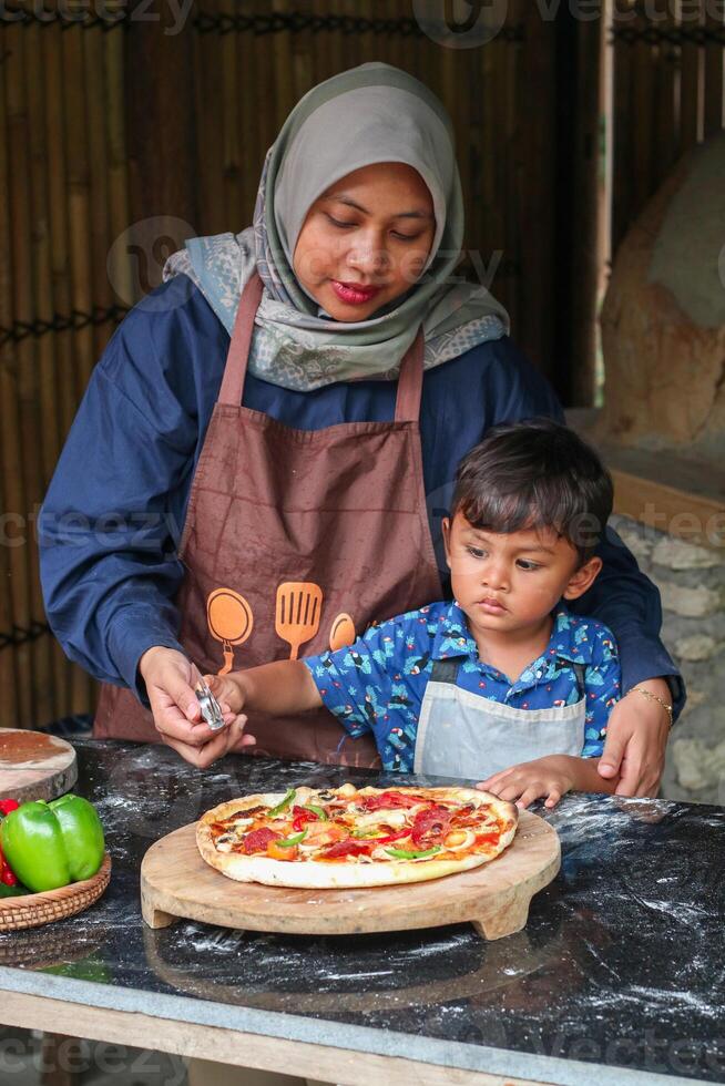 Asian Muslim Mother And Little son Rolling Up Dough On Table While Baking In Kitchen Together, Cute Small Girl And Islamic Mom In Hijab Having Fun At Home, Enjoying Preparing Homemade Pastry photo