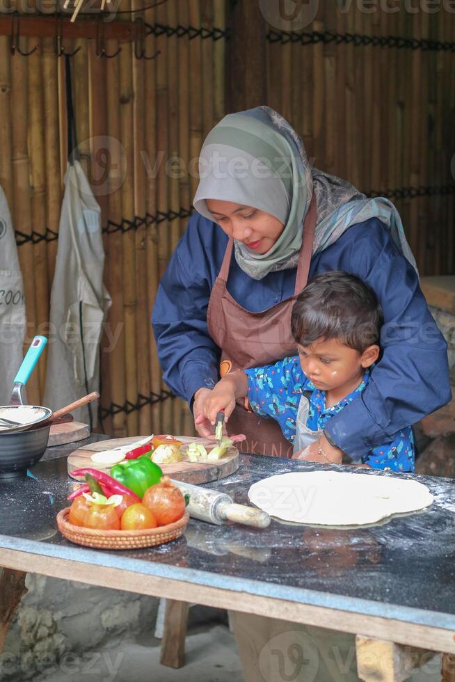 Happy family adult parent hijab mom teaching small child son doing bakery, cute little kid boy hold rolling pin roll dough having fun helping mum learning bake pastry make pie or biscuits in kitchen photo