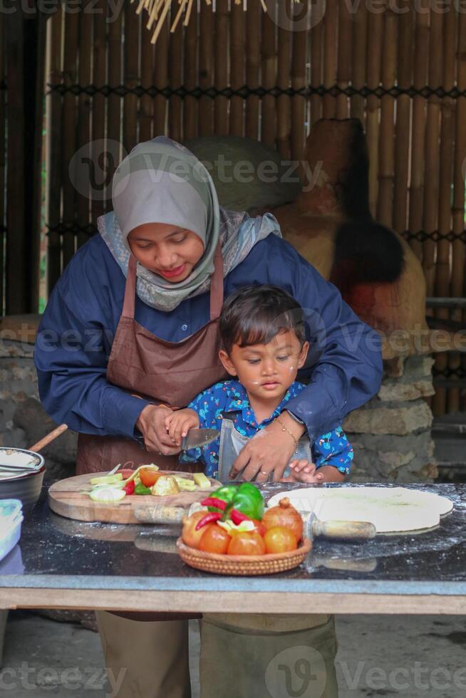 Happy family adult parent hijab mom teaching small child son doing bakery, cute little kid boy hold rolling pin roll dough having fun helping mum learning bake pastry make pie or biscuits in kitchen photo