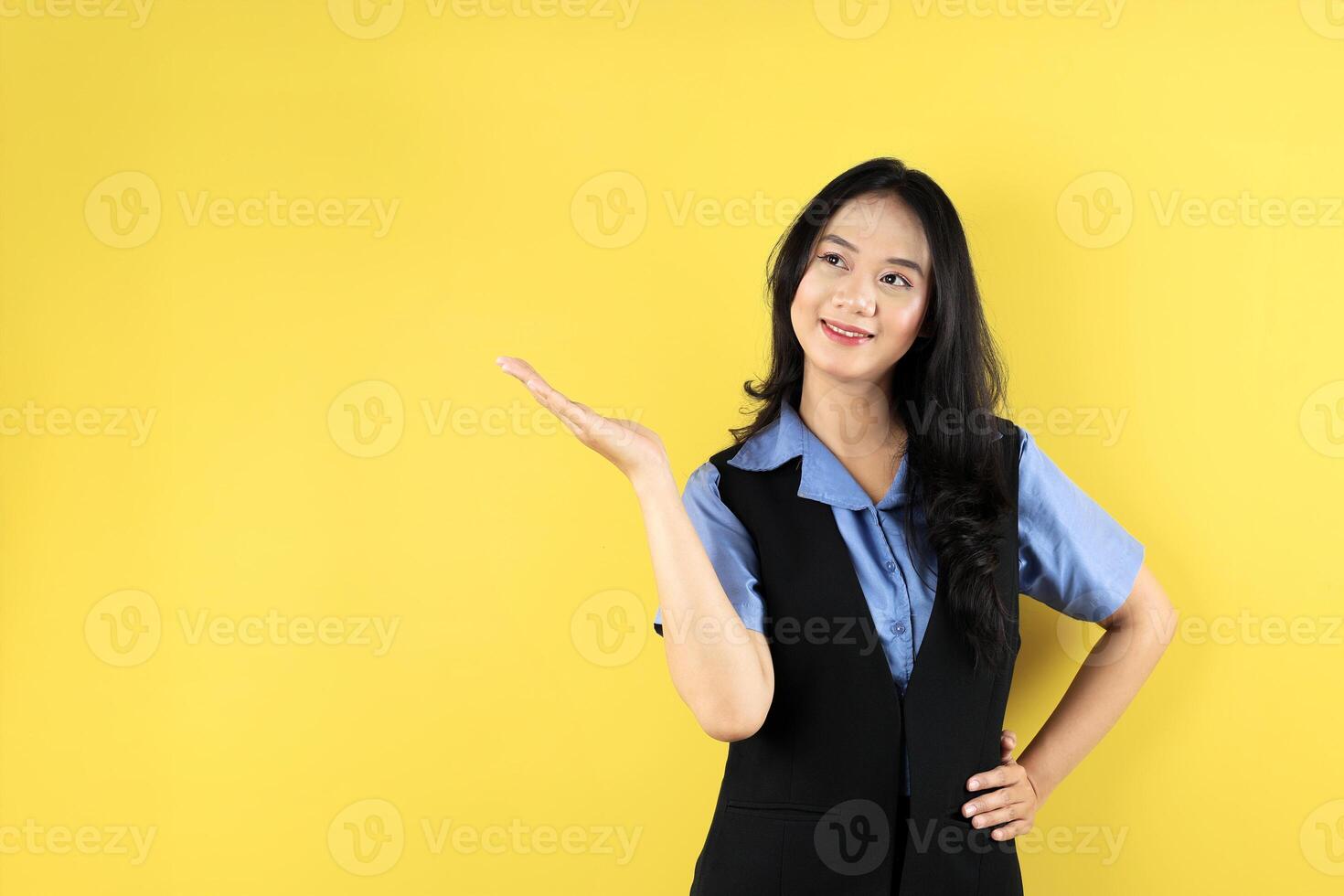 Asian Woman Wearing Blue Shirt Pointing to an Empty Space in a Studio with Yellow Background. photo