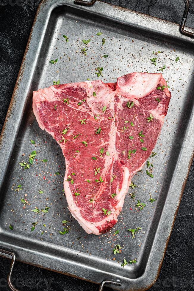 T-bone steak, raw marbled beef meat in a steel kitchen tray with herbs. Black background. Top view photo