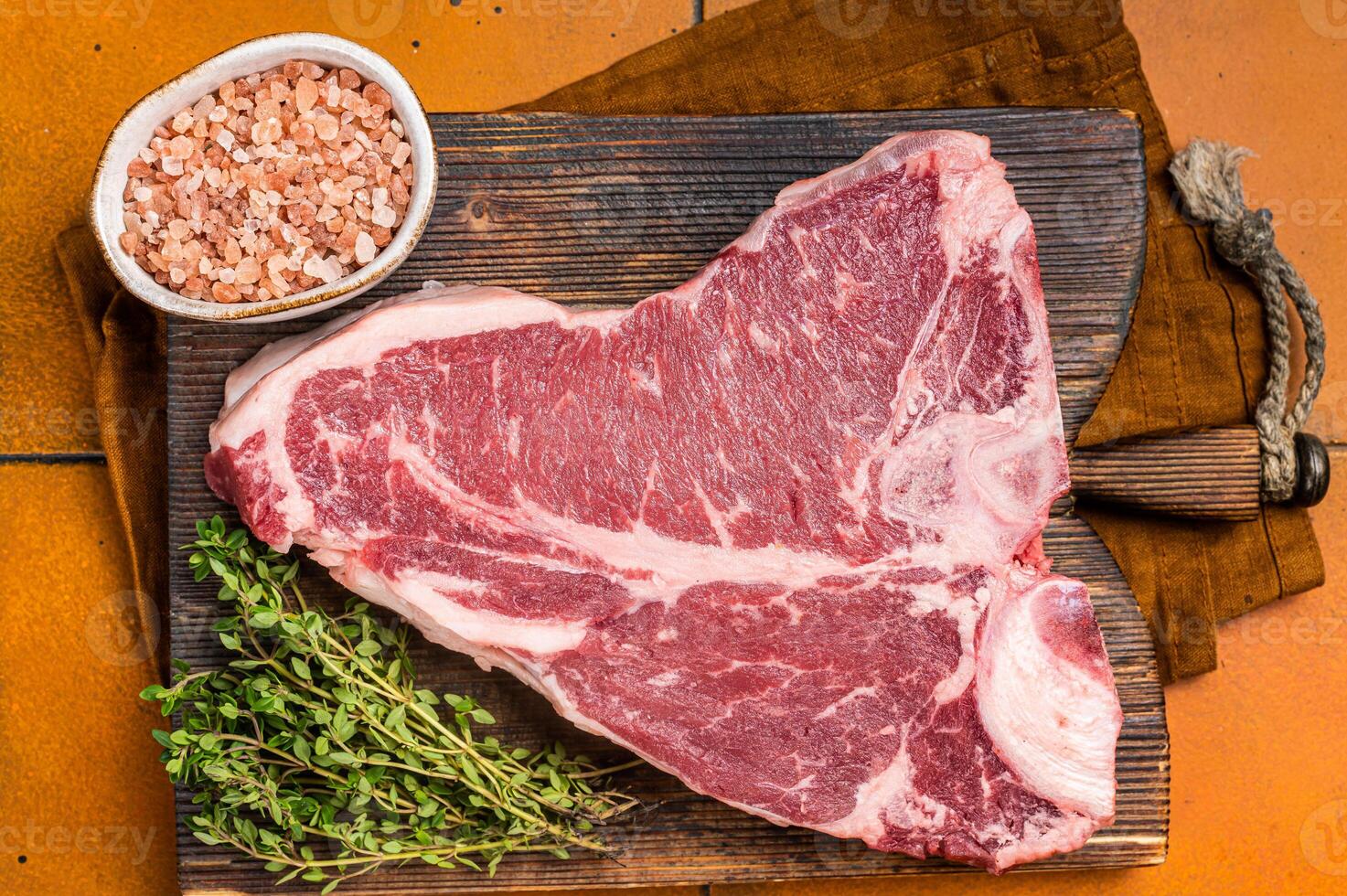 Porterhouse raw beef meat Steak, T-bone with thyme and sea salt on a wooden board. Orange background. Top view photo