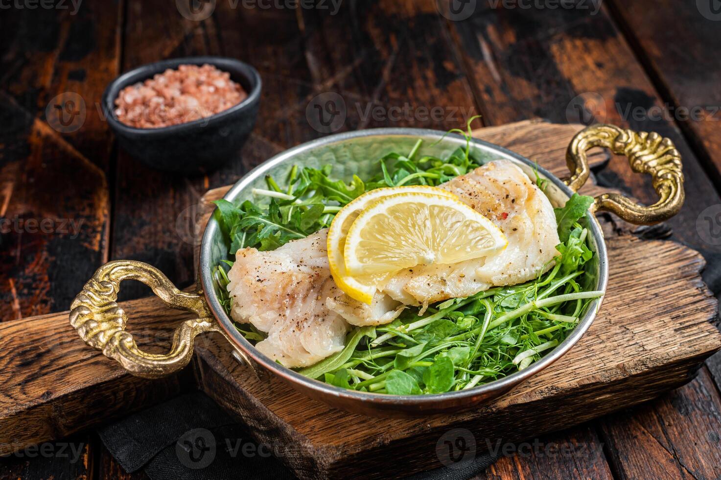 Roasted pollock or coalfish fish fillet served with green salad in a skillet. Wooden background. Top view photo