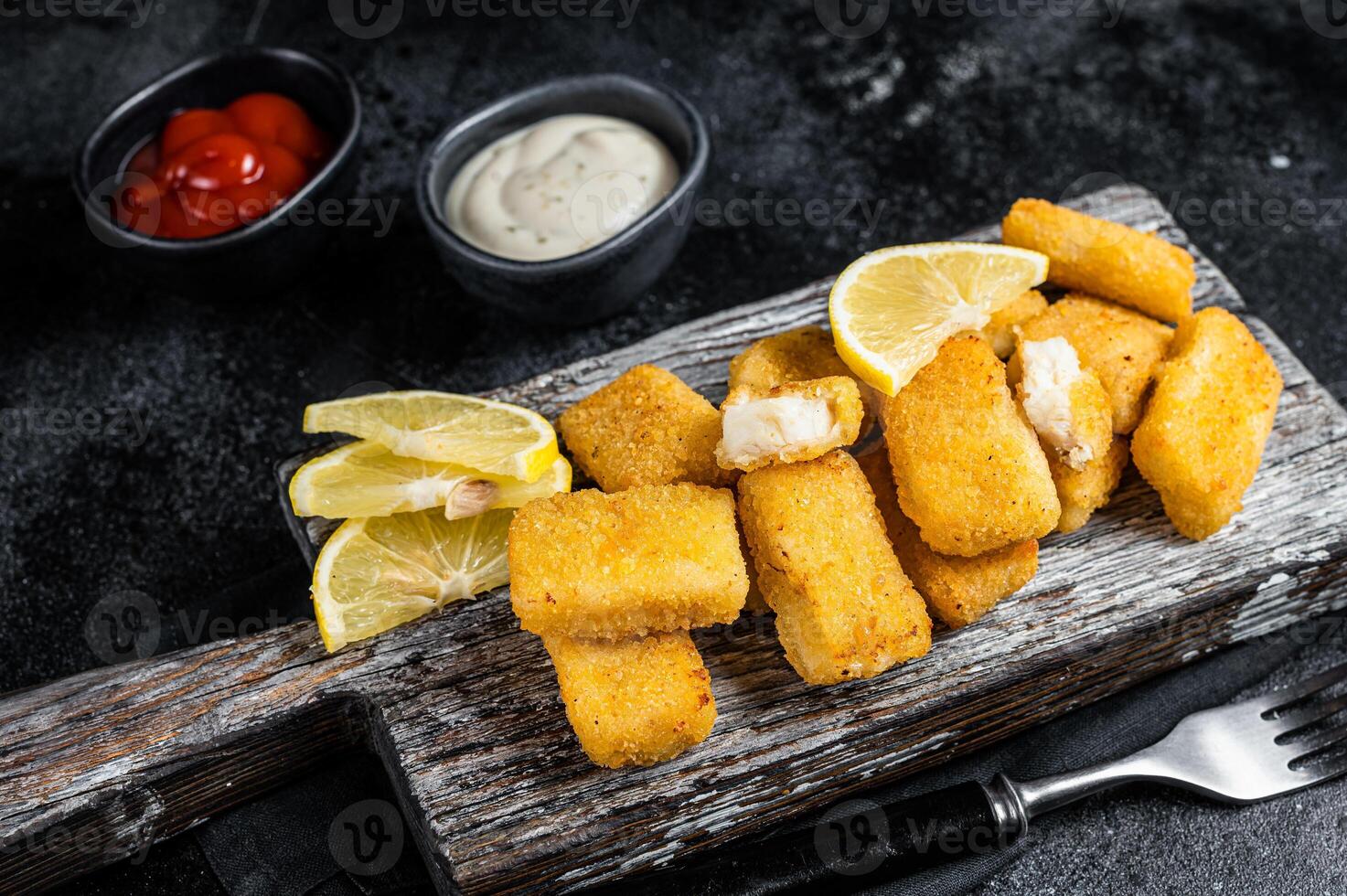 Golden roast Fish Sticks Fingers with sauce on a wooden board. Black background. Top view photo