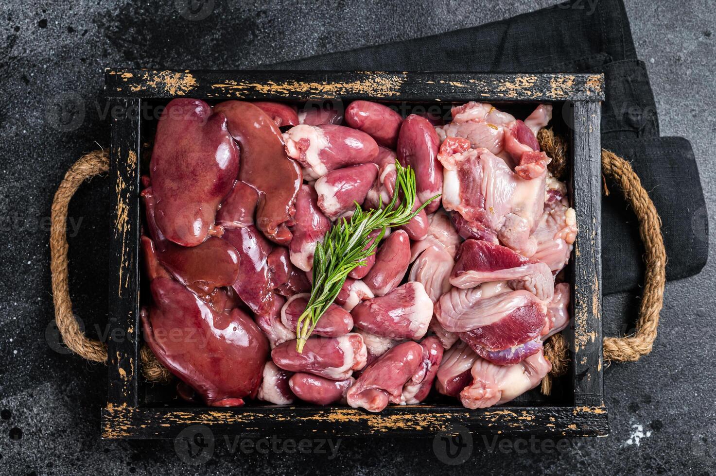 Uncooked Bird offals, raw chicken giblets, gizzards, stomachs, liver and hearts in a wooden tray. Black background. Top view photo