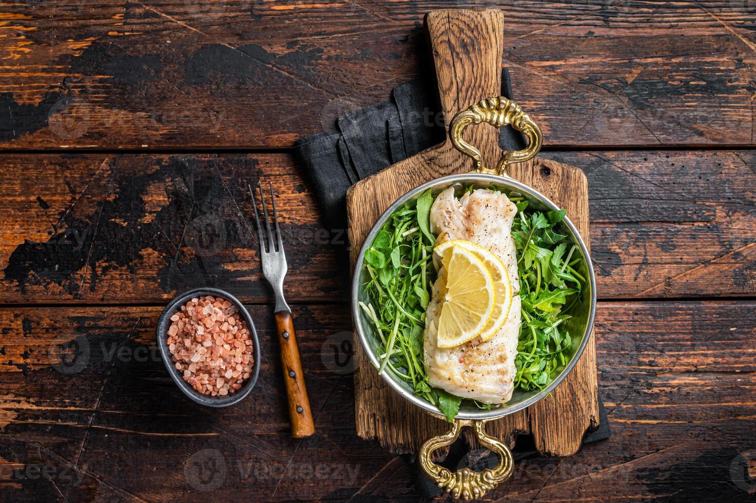 Roasted pollock or coalfish fish fillet served with green salad in a skillet. Wooden background. Top view photo