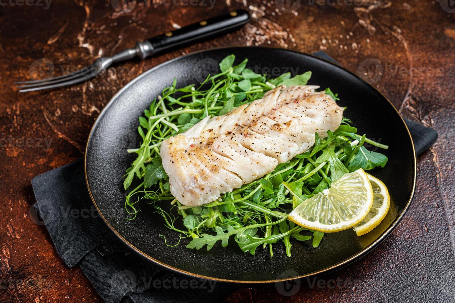 Grilled cod fish fillet served with green salad in a plate. Dark background. Top view photo