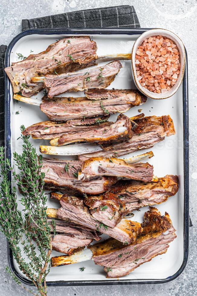 BBQ smoked lamb ribs, mutton spareribs, sliced meat in steel tray with herbs. Gray background. Top view photo