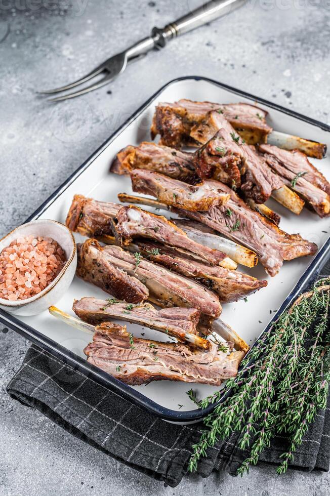 BBQ smoked lamb ribs, mutton spareribs, sliced meat in steel tray with herbs. Gray background. Top view photo