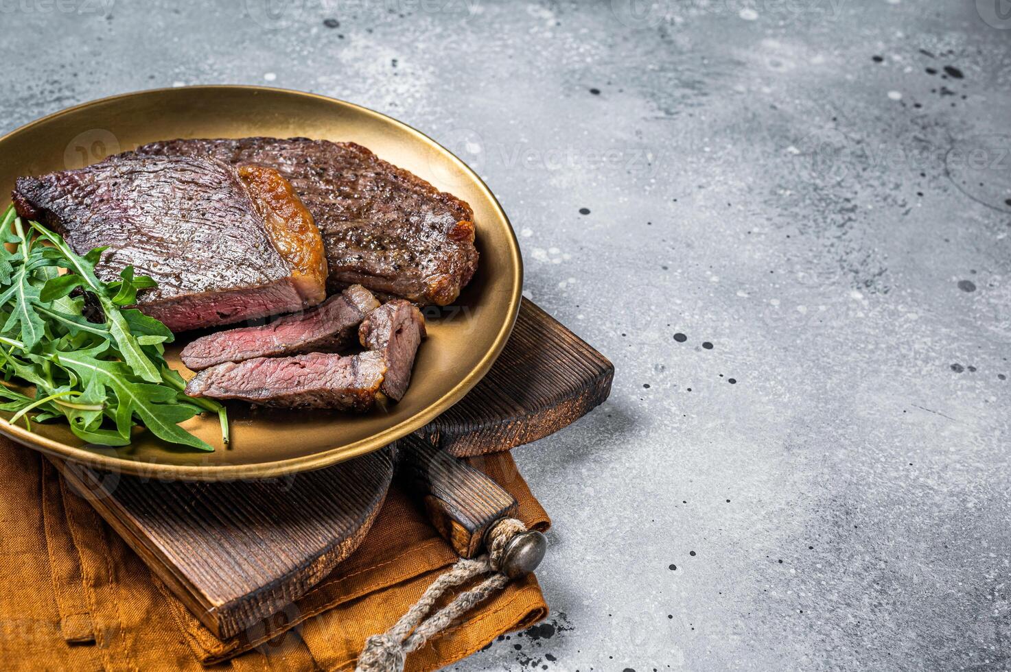 Roast picanha or top sirloin beef meat steak on a plate with arugula salad. Gray background. Top view. Copy space photo