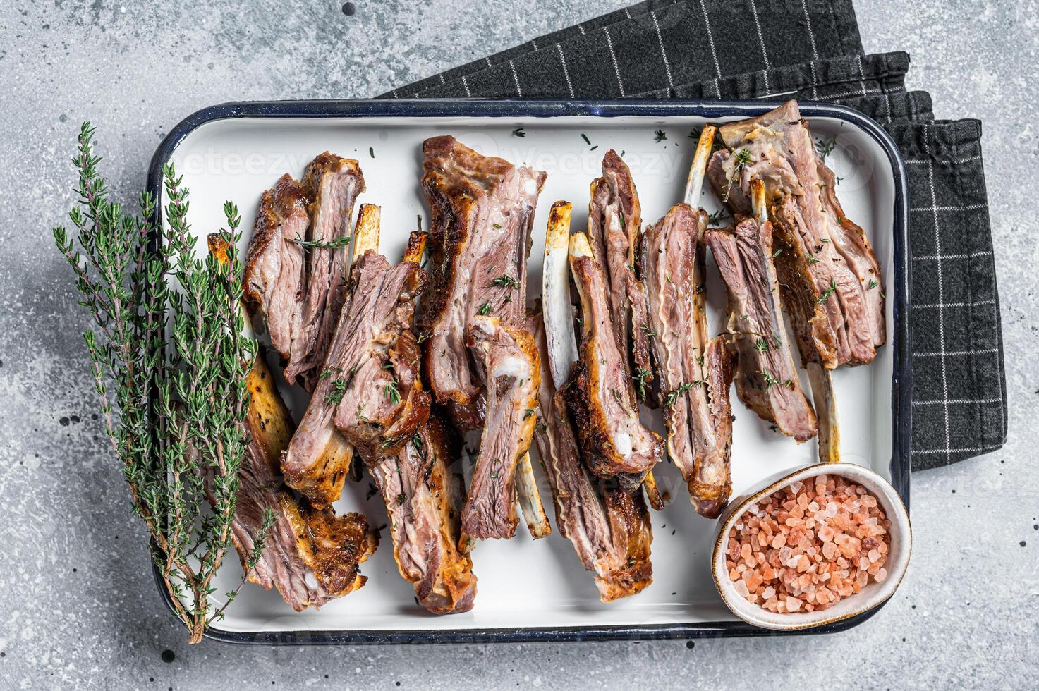 BBQ smoked lamb ribs, mutton spareribs, sliced meat in steel tray with herbs. Gray background. Top view photo