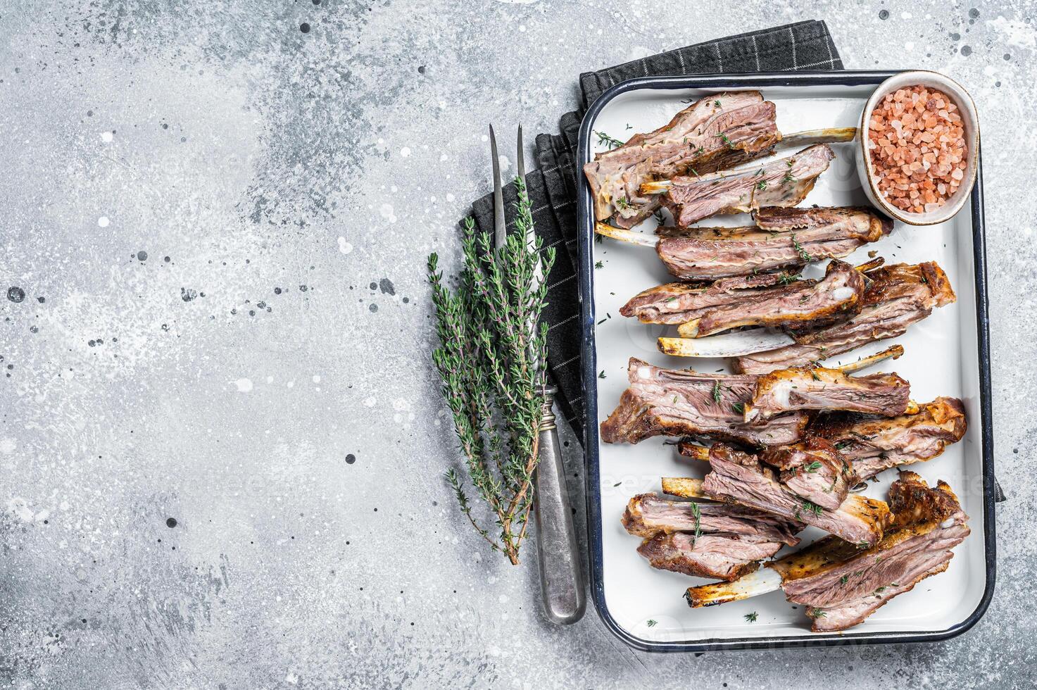 BBQ smoked lamb ribs, mutton spareribs, sliced meat in steel tray with herbs. Gray background. Top view. Copy space photo