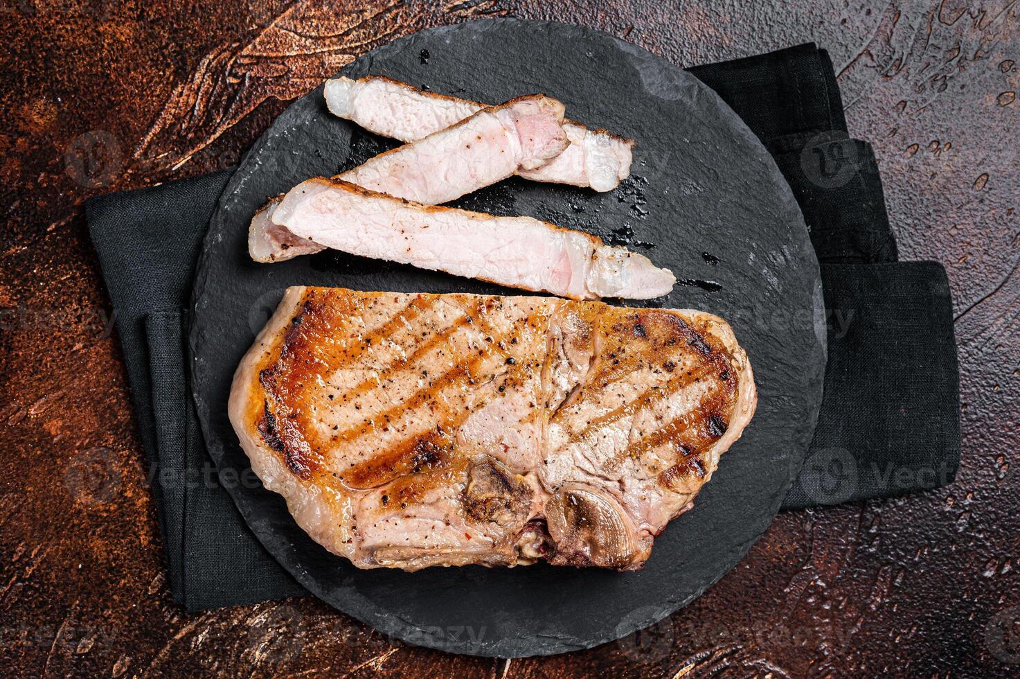 Roast BBQ pork Chop or T-bone meat steak sliced on a board. Dark background. Top view photo
