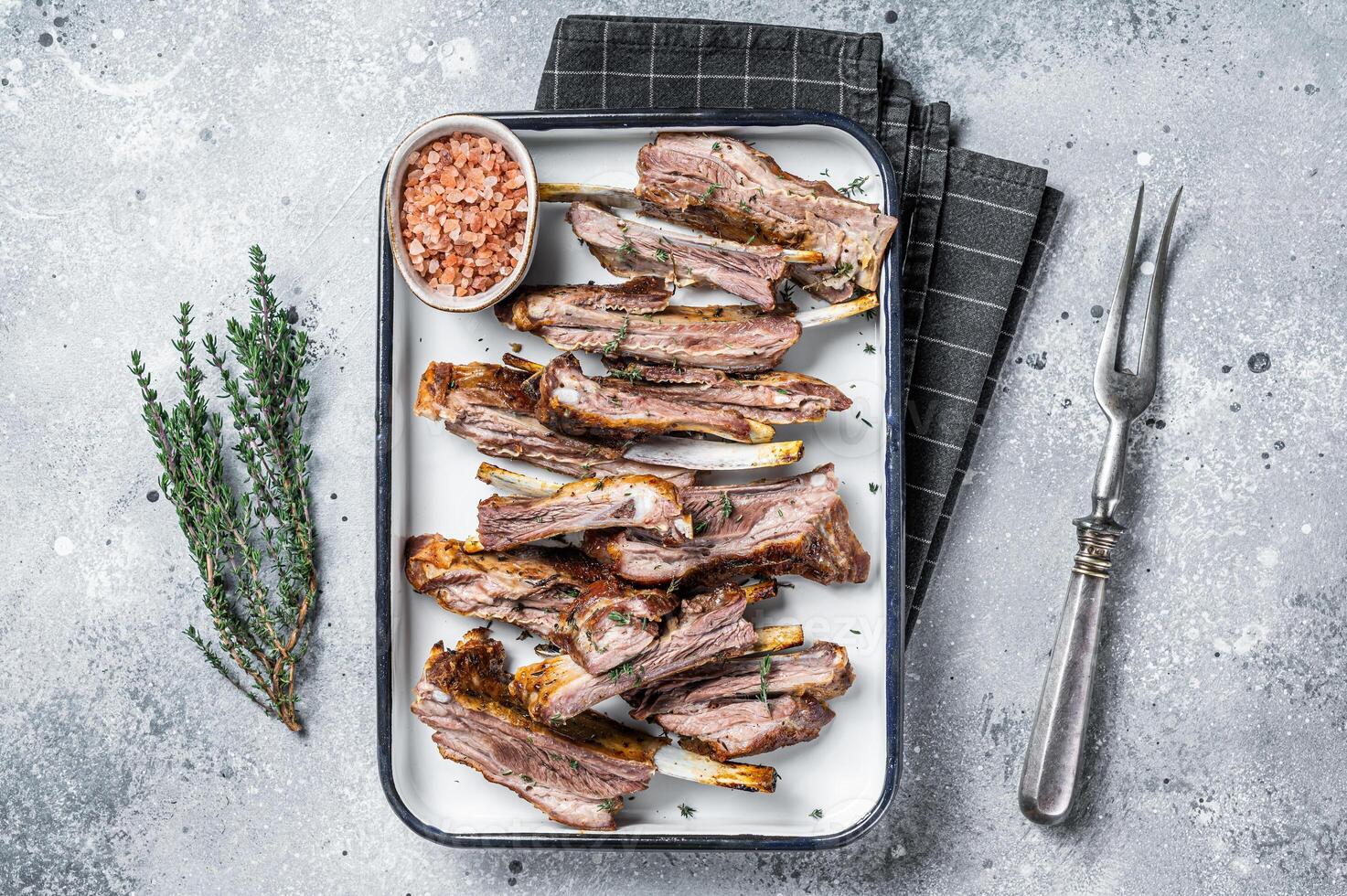 BBQ smoked lamb ribs, mutton spareribs, sliced meat in steel tray with herbs. Gray background. Top view photo