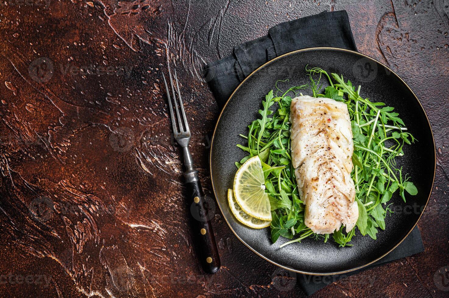 Grilled cod fish fillet served with green salad in a plate. Dark background. Top view. Copy space photo