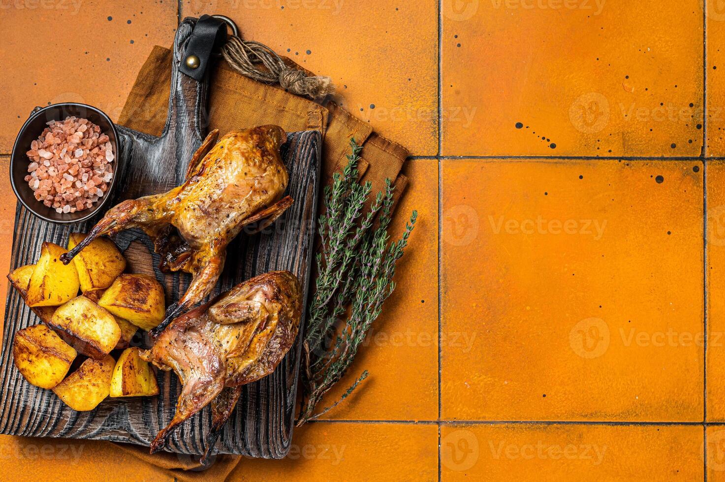Roasted quails with potatoes, spices and herbs on a wooden board. Orange background. Top view. Copy space photo