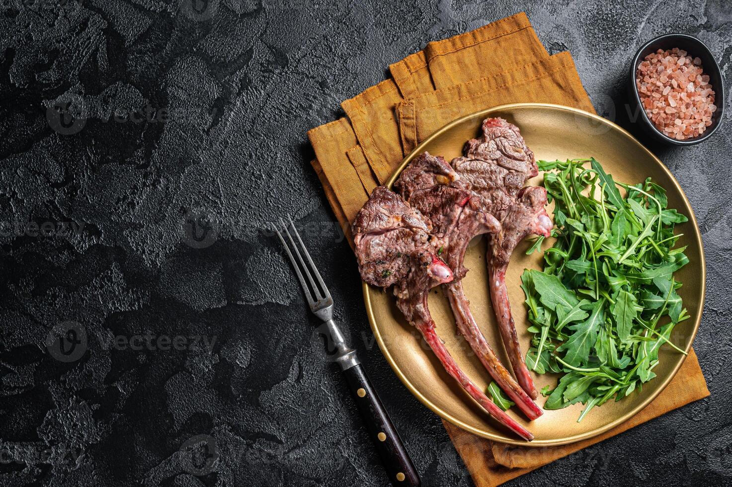 BBQ grilled lamb cutlet or mutton chop steak with arugula salad on a plate. Black background. Top view. Copy space photo