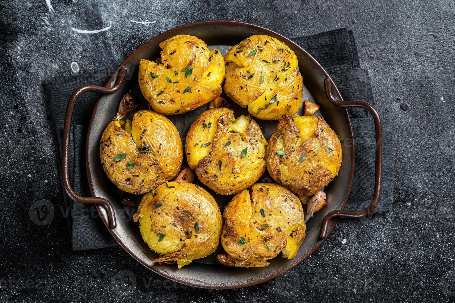 Broken potatoes baked in oil with herbs. Black background. Top view photo
