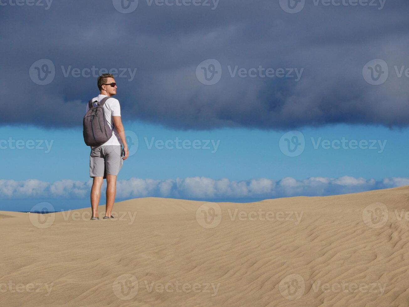 A man traveler tourist wearing sunglasses and backpack walks through the desert at the background of cloudy sky. Dune in Maspalomas, Gran Canaria, Spain photo