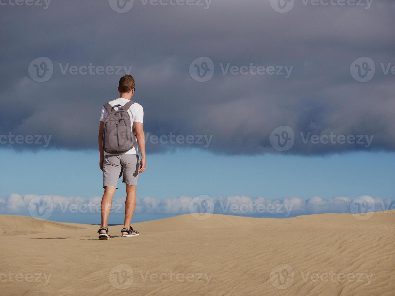 A man traveler tourist with a backpack walks through the desert at the background of cloudy sky. Dune in Maspalomas, Gran Canaria, Spain photo
