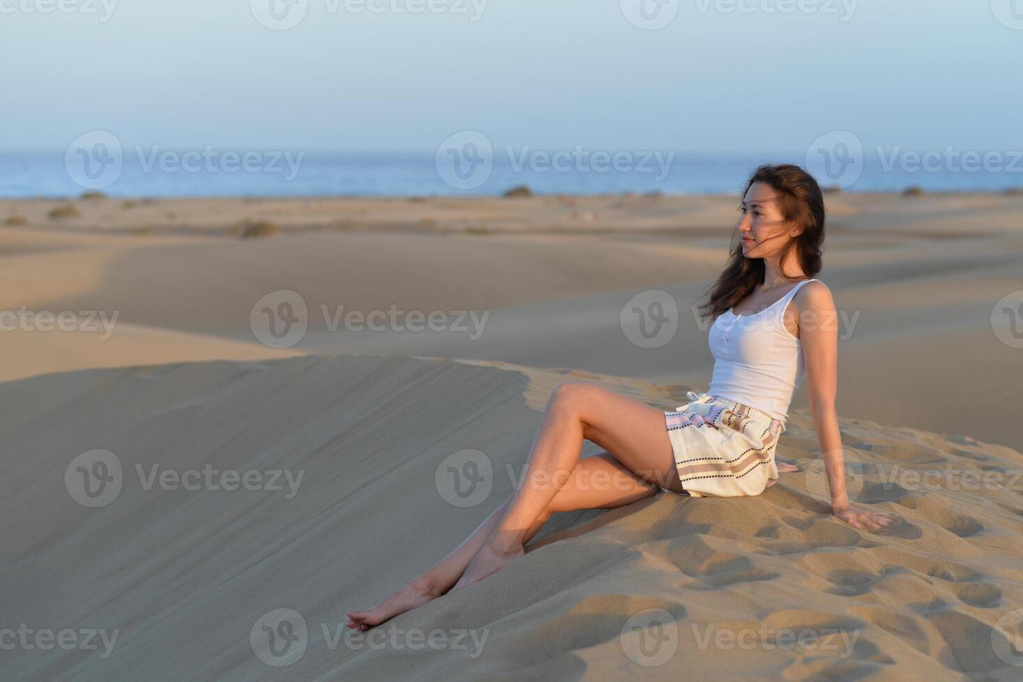 A girl seating on a sand at the background of a sea at a golden hour before sunset. Dune in Maspalomas, Gran Canaria. Touristic travel concept photo
