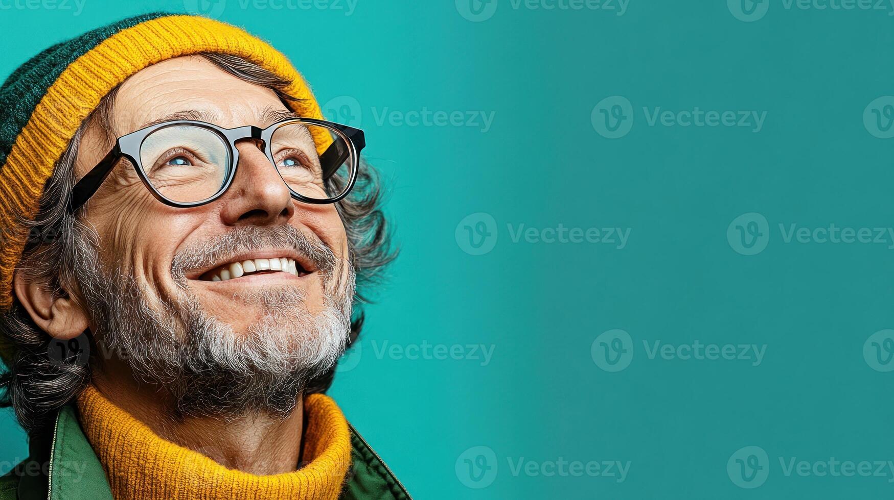 An older man beams with happiness, wearing glasses and a colorful knitted hat, set against a bright turquoise backdrop photo