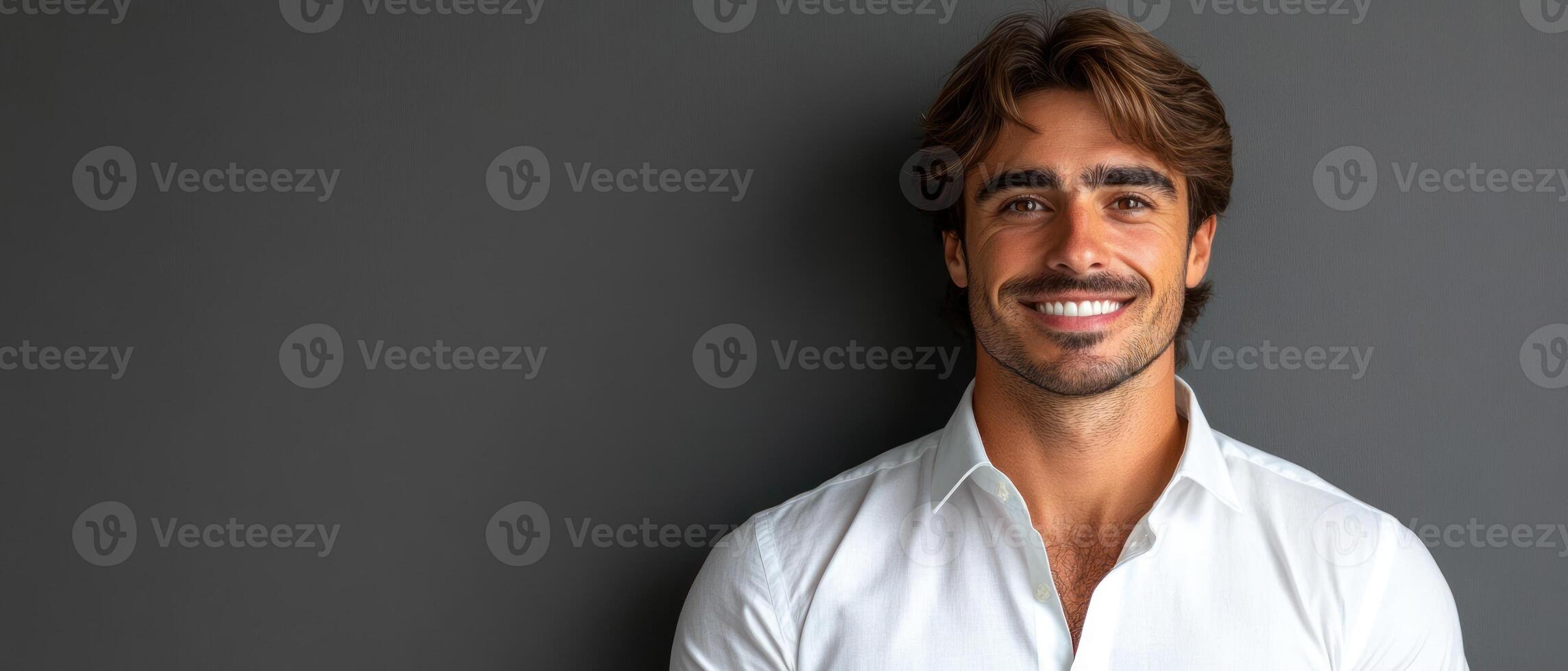 A cheerful man in a white shirt stands confidently against a sleek dark wall, sporting a genuine smile photo