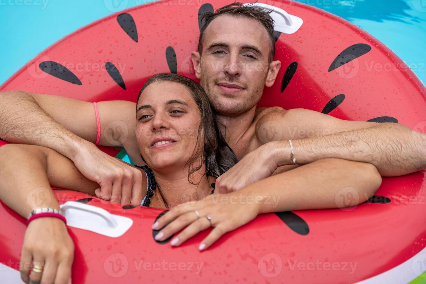 young couple in swimsuits having fun in the splashing blue water of a swimming pool inside a red inflatable life ring photo