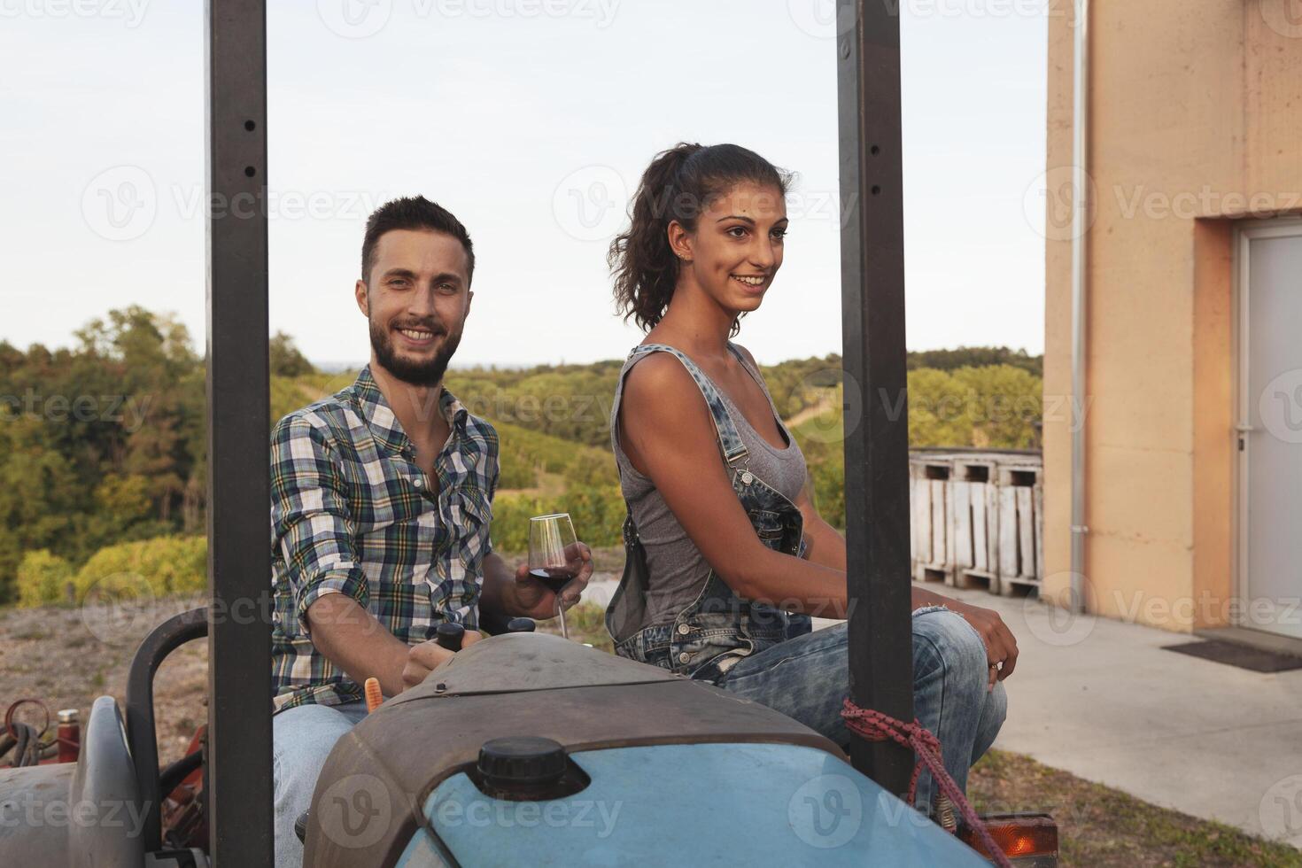 young couple of winemakers toasting with two glasses of wine sitting on an old crawler tractor at sunset photo