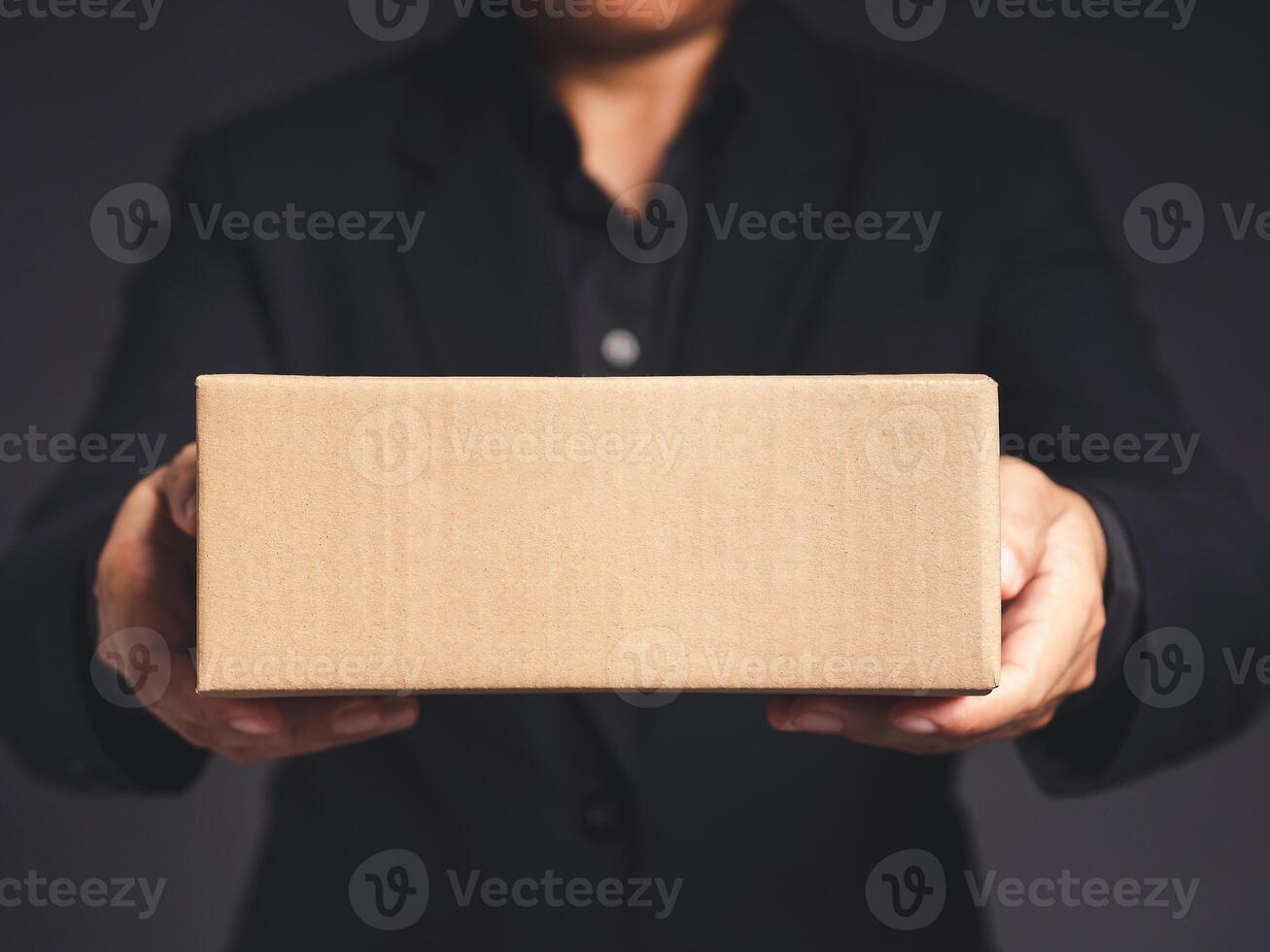 Businessman in a black suit holds and points to a cardboard box while standing on a gray background photo